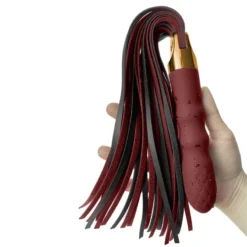 Mão segurando o vibrador chicote pelo cabo com as tiras bordô e pretas caindo